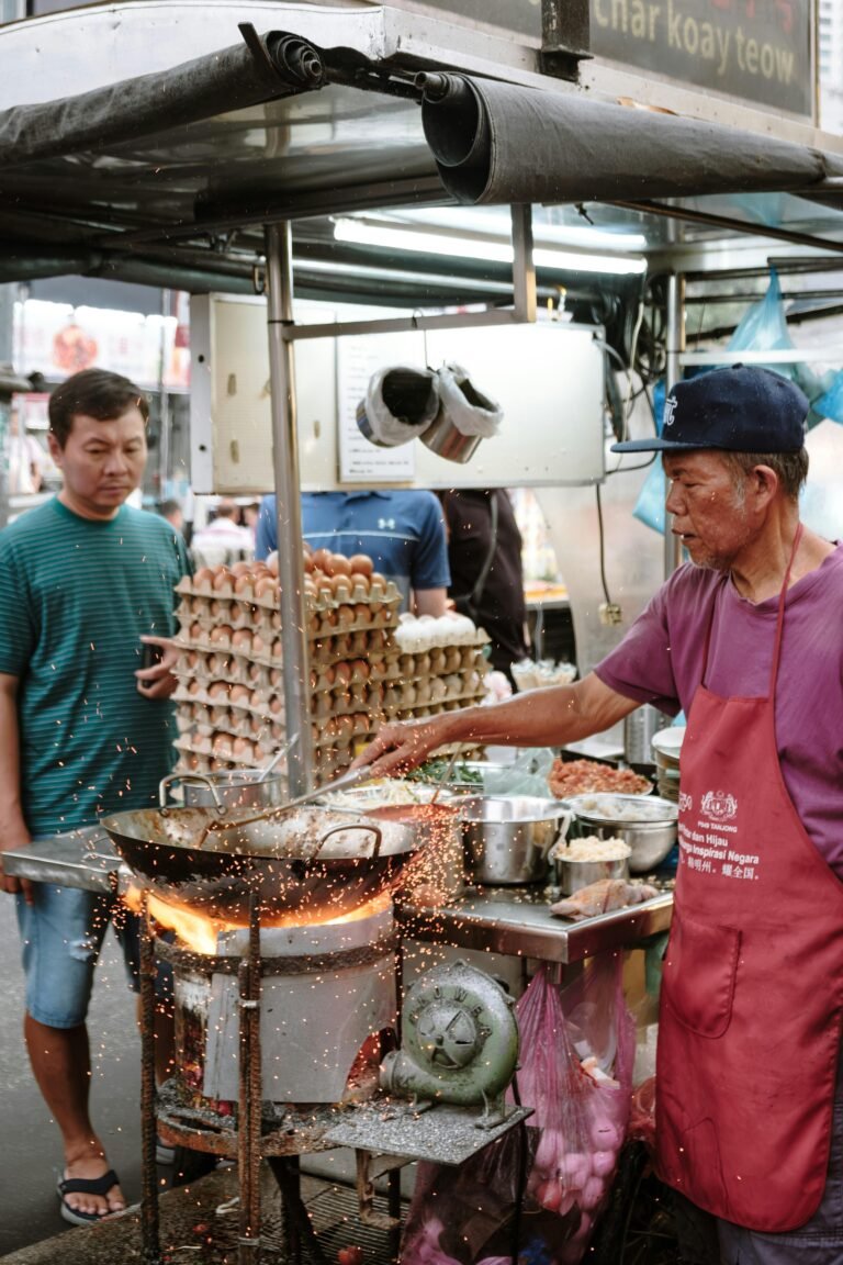 Street vendor preparing char kway teow with sparks flying from a wok in an outdoor market.